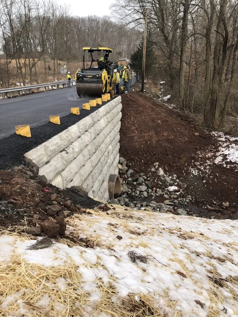 An image of PennDOT workers in yellow safety apparel and white hard hats working with yellow PennDOT construction equipment on top of the newly reconstructed roadway above the new retaining wall.